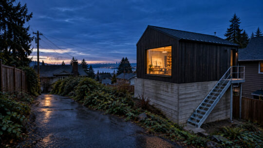 A charred cedar studio standing on a board-formed concrete base that retains eight feet of grade, seen from the alley.
