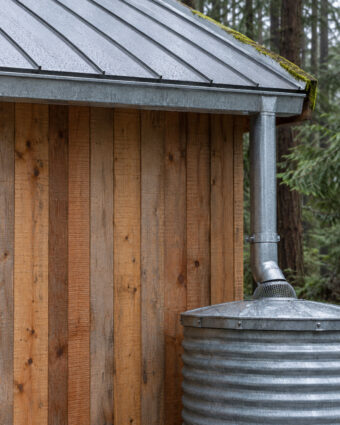 A standing-seam roof edge draining through a single galvanized downspout into a corrugated steel cistern.